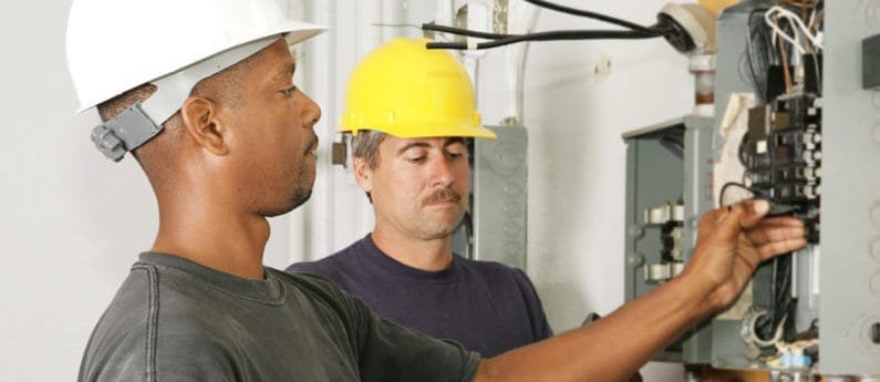 Two HVAC technicians in hard hats work on a panel. 